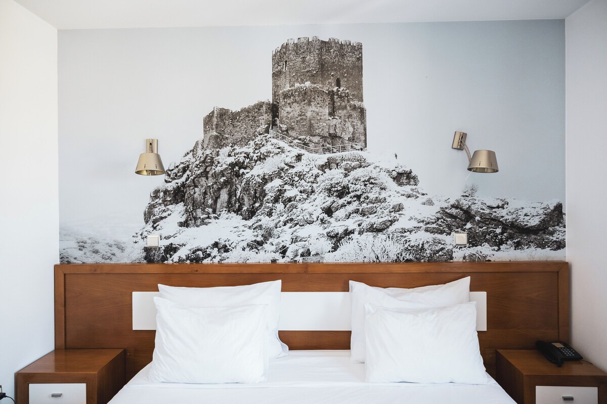A hotel room with a large bed, wooden headboard, two bedside tables, and a wall-mounted lamp.