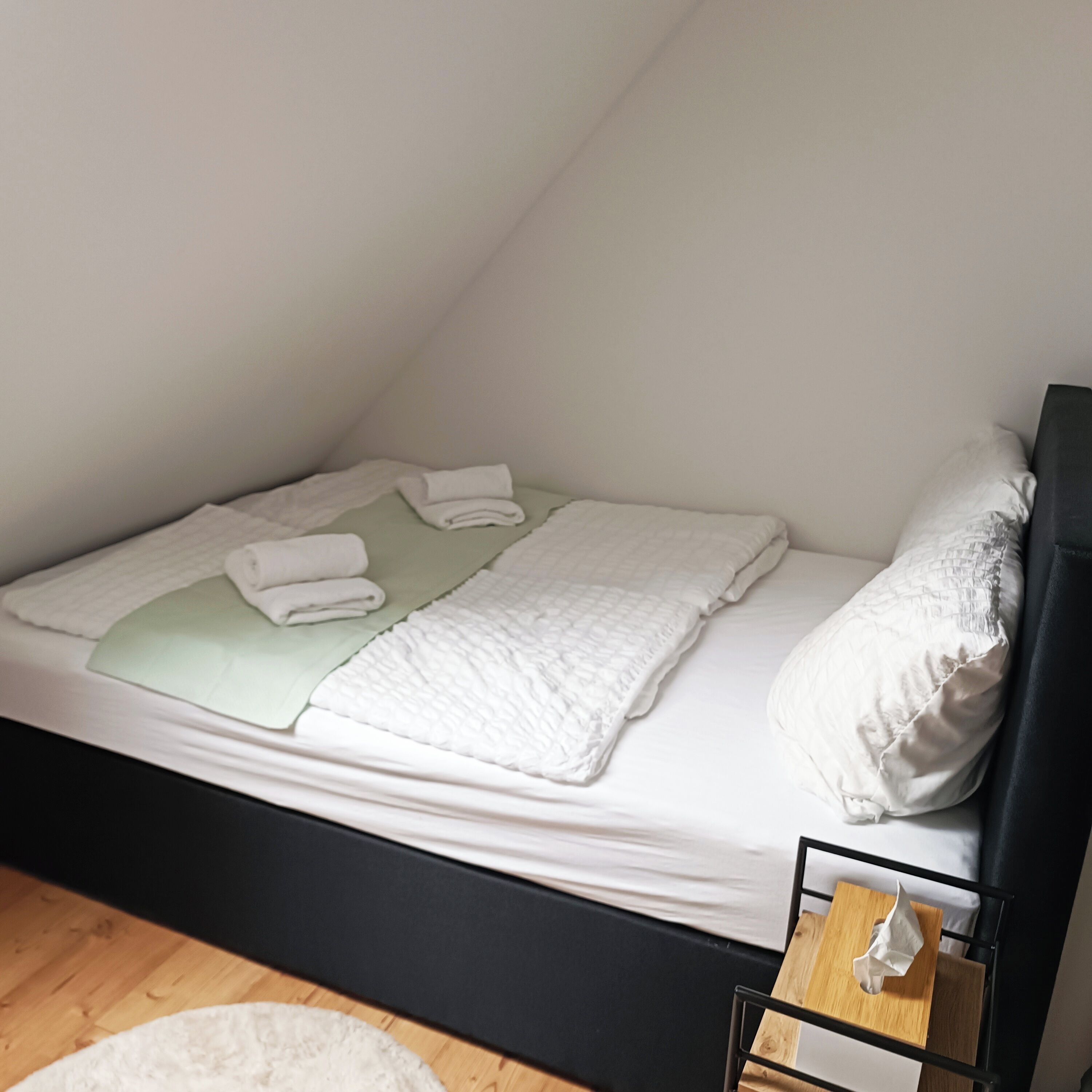 Una habitación pequeña con cama individual, suelo de madera, cabecera negra y mesita de noche con caja de pañuelos.