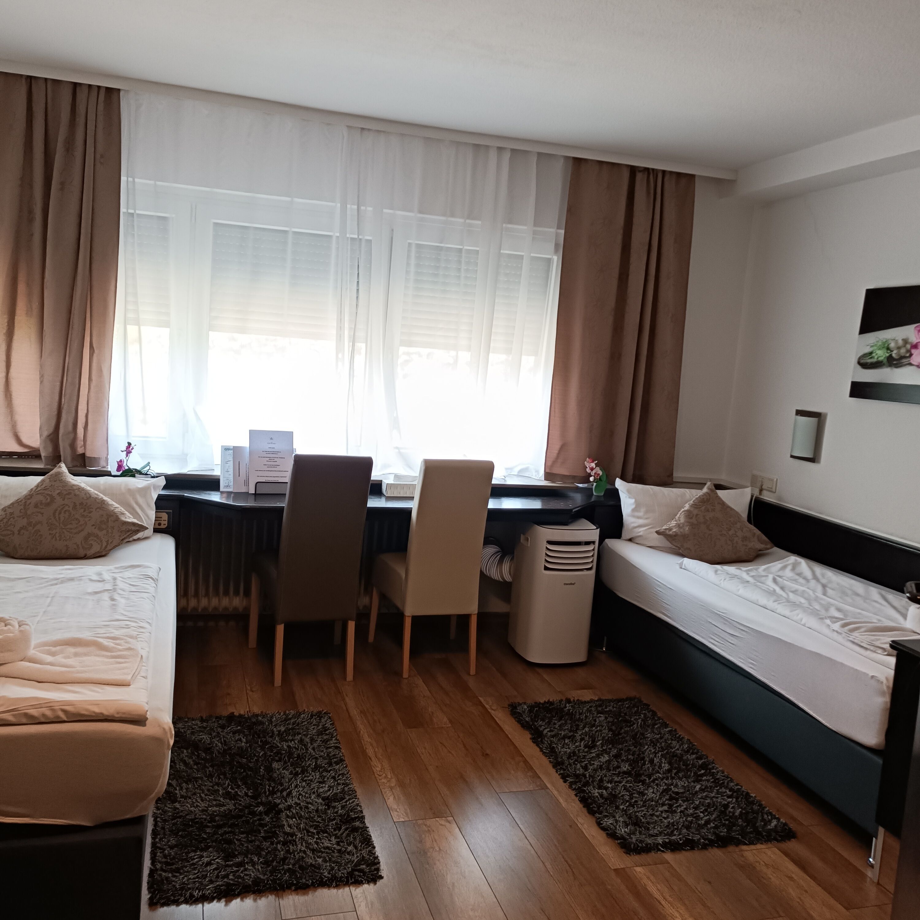 Quarto de hotel com cama, escrivaninha, cadeira, radiador e janela com cortinas.