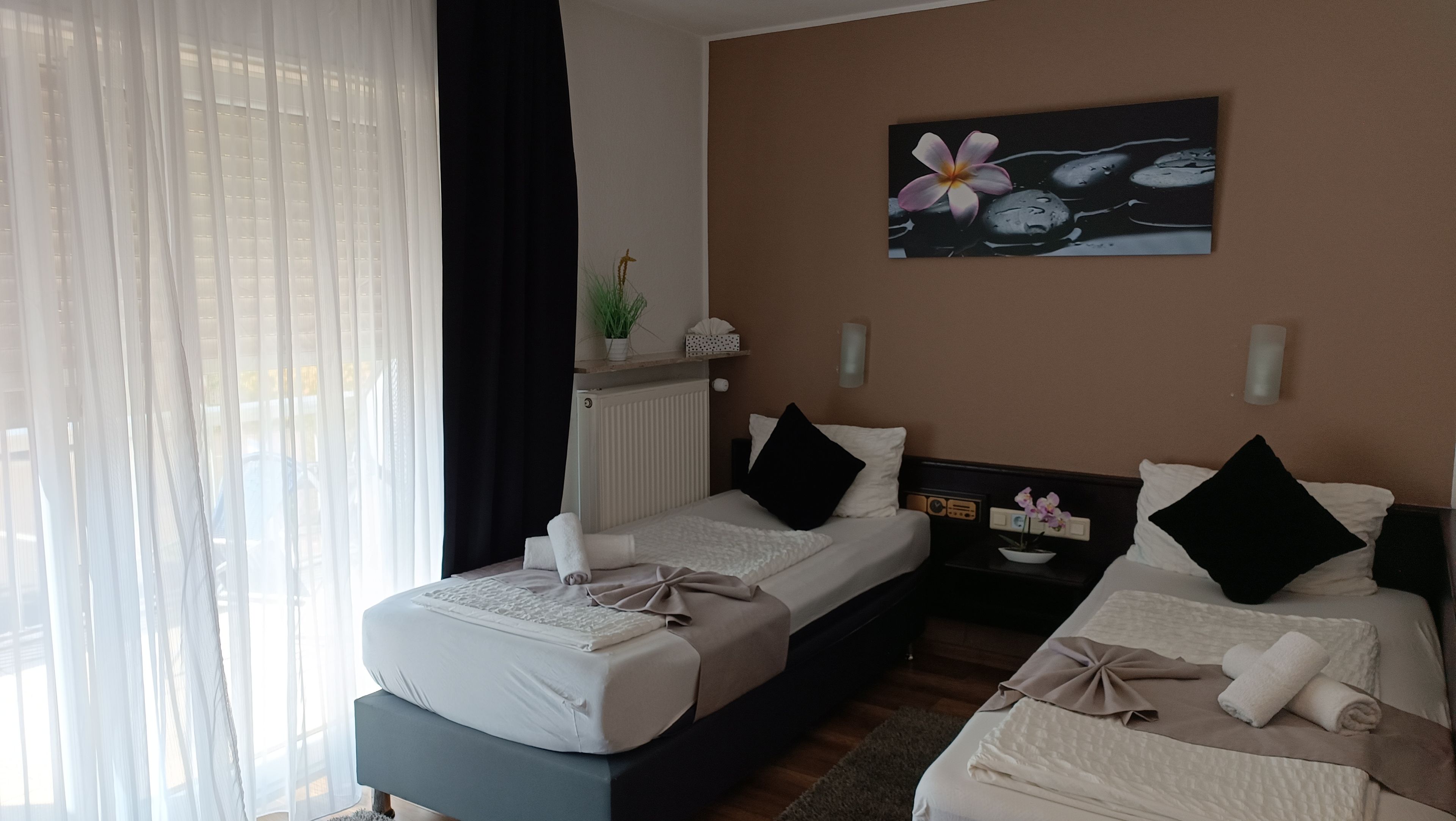 A hotel room with two beds, a window with sheer curtains, a painting on the wall, and a small table with a plant.
