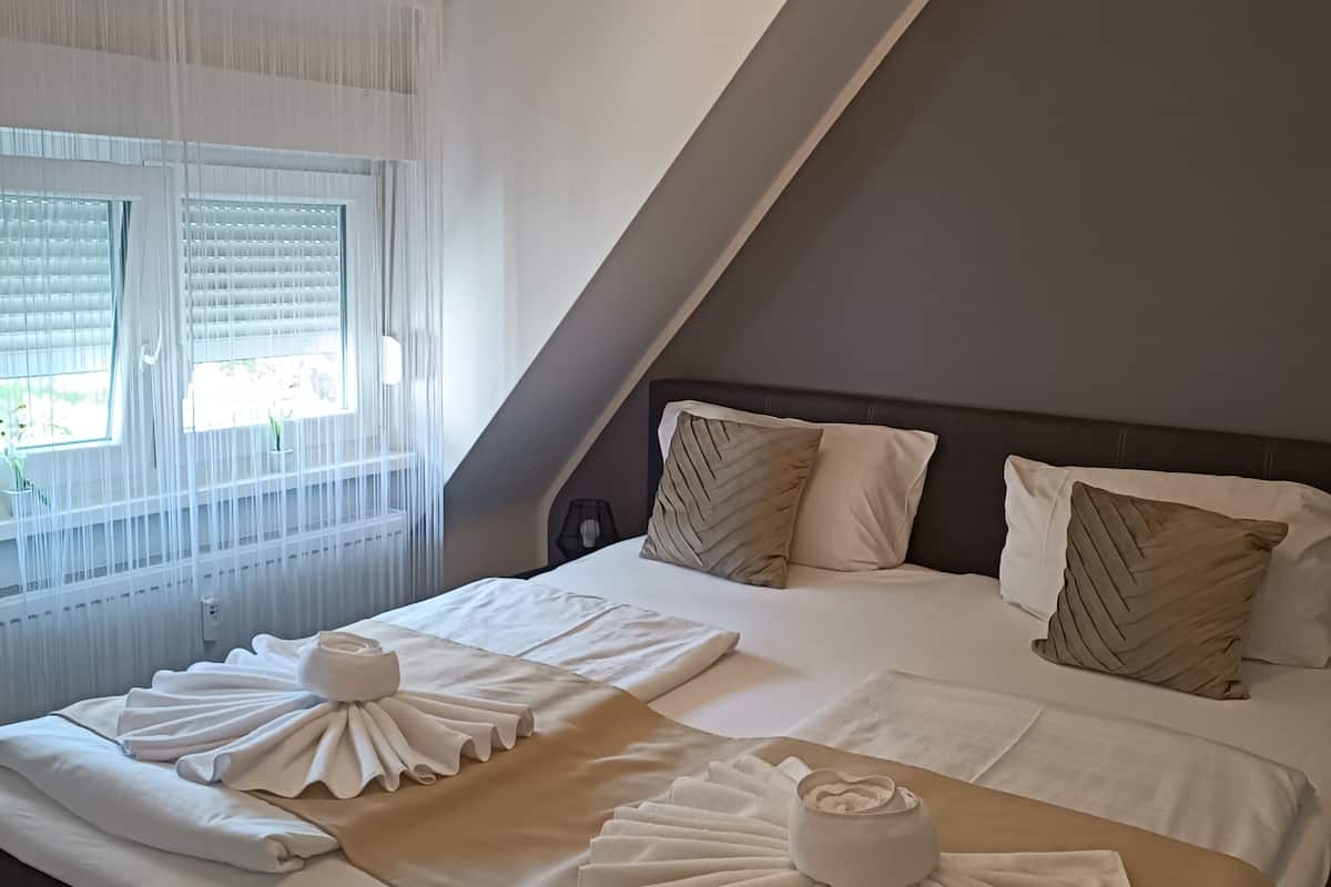 Une chambre d’hôtel avec un lit, deux oreillers, une table de chevet et une fenêtre avec des rideaux légers.
