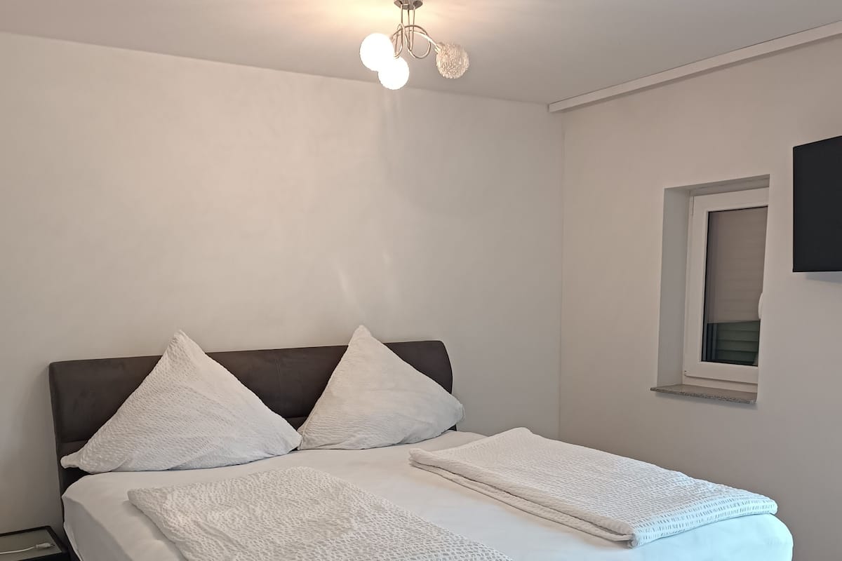 Un lit bien fait, avec du linge de lit blanc et deux oreillers, une tête de lit sombre et une petite table de chevet équipée d’une prise électrique.