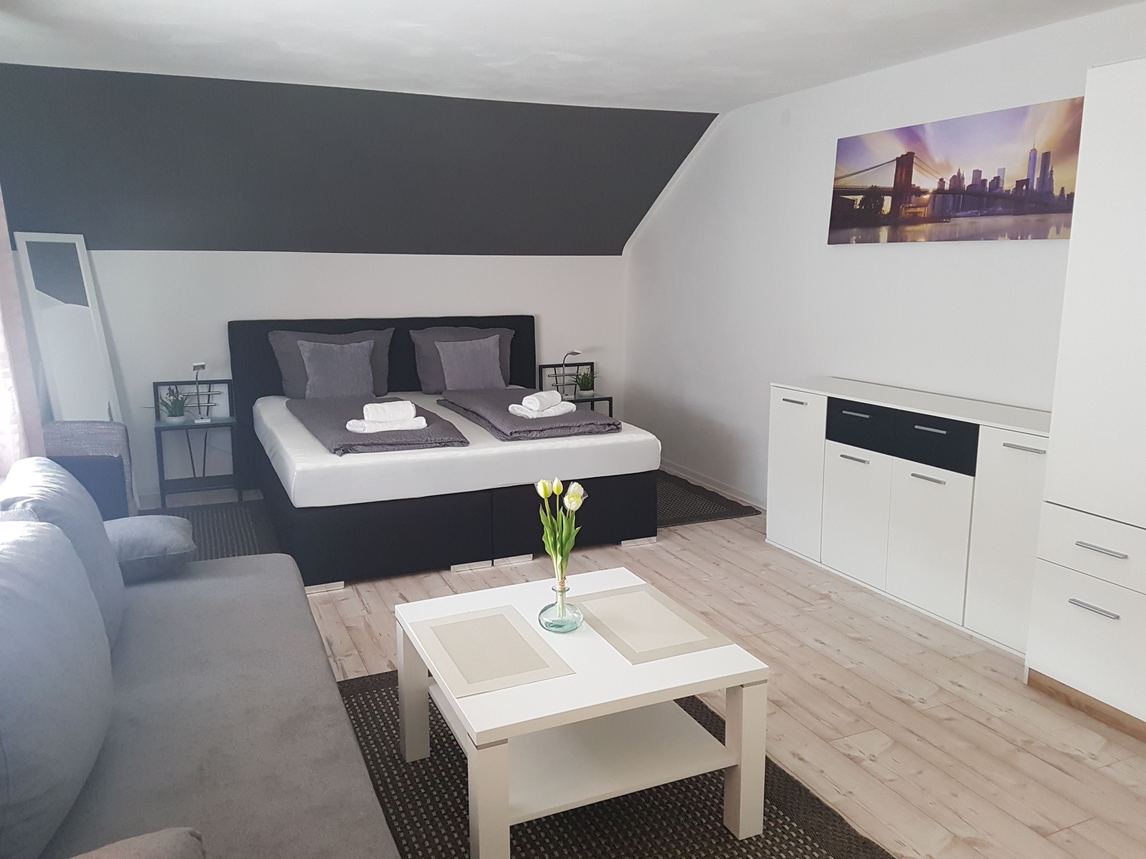 Un dormitorio moderno con cama, mesitas de noche, una pequeña mesa con un florero, un sofá y una kitchenette.