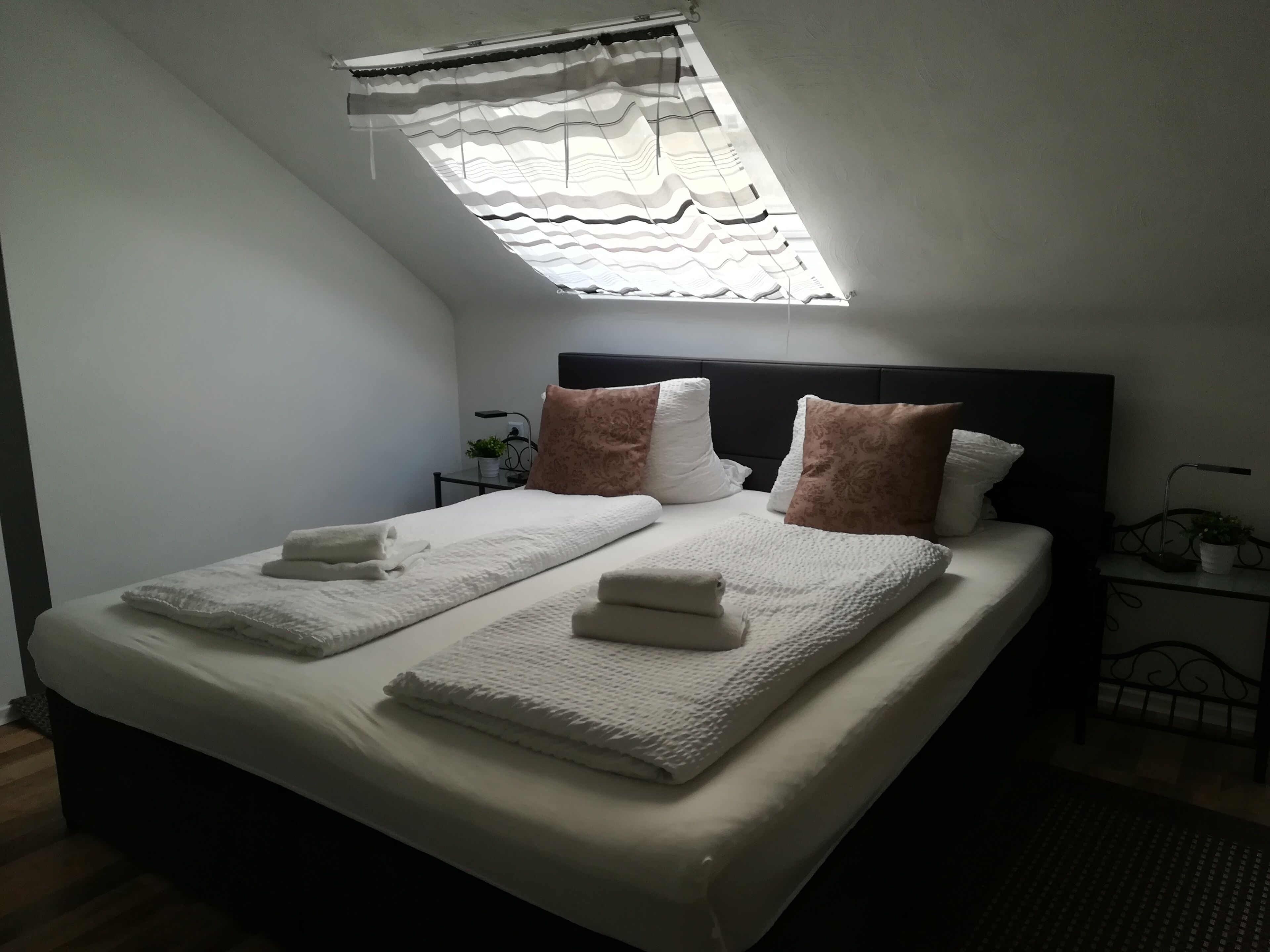 Un lit bien fait, avec deux oreillers et deux serviettes pliées, une table de chevet avec une lampe et une fenêtre ornée d’un rideau à motifs.
