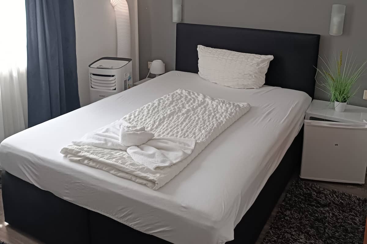 Un lit bien fait, avec du linge de lit blanc, une tête de lit sombre et une table de chevet sur laquelle se trouvent une lampe et une plante.