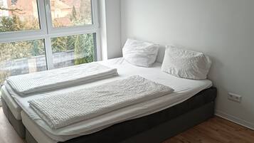 Ein Schlafzimmer mit großem Fenster, einem Bett mit weißer Bettwäsche und einem grauen Matratze.