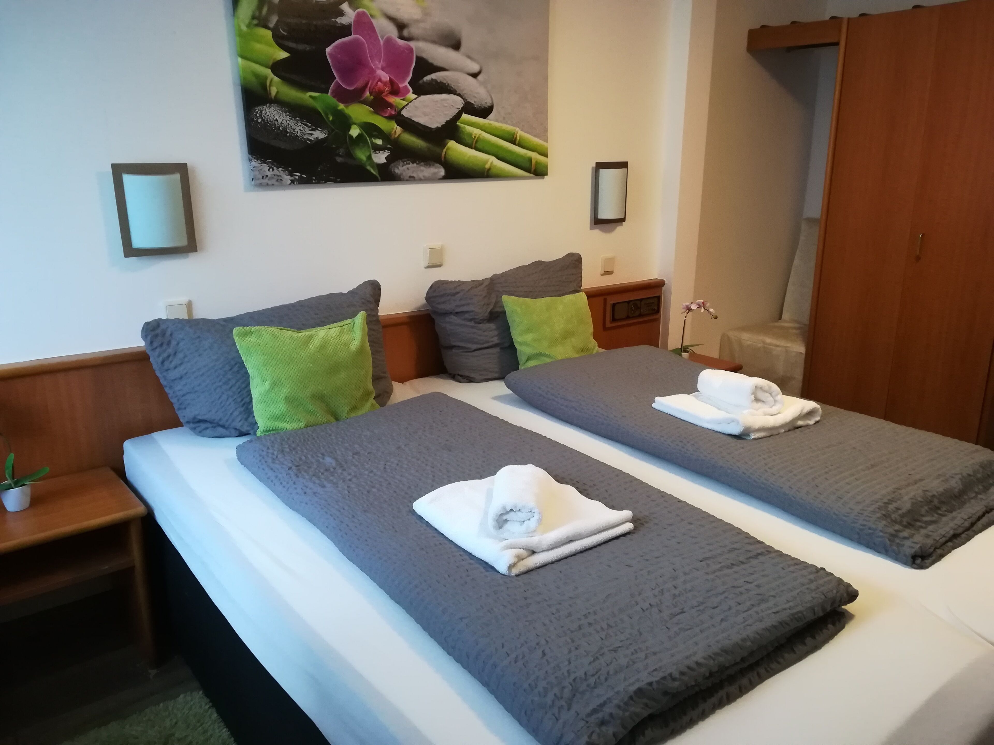 A hotel room with two beds, grey and white bedding, green and grey pillows, a small bedside table with a plant, and a wall-mounted picture of flowers and rocks.