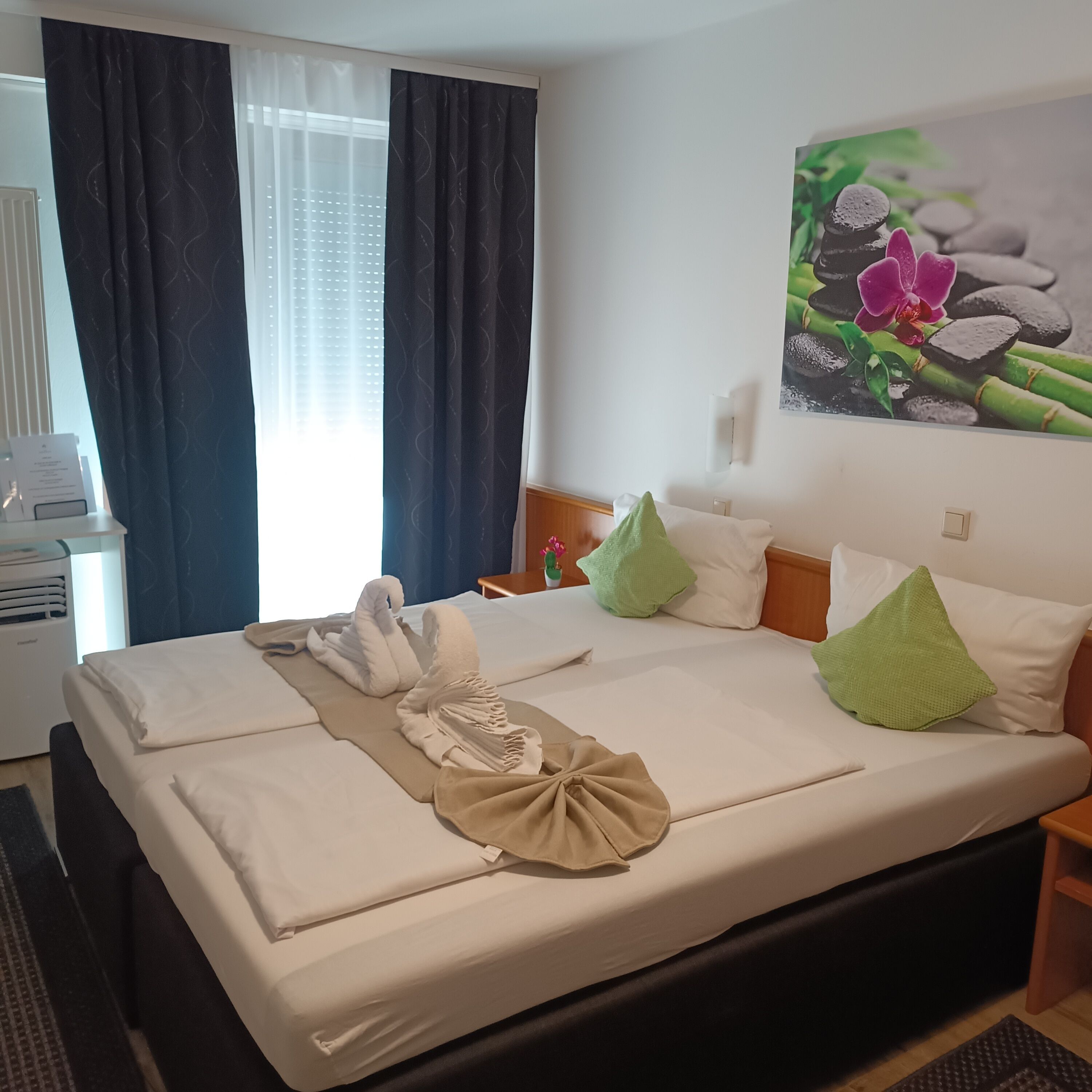 A hotel room with a bed, a nightstand, a wall-mounted lamp, a window with curtains, and a painting of flowers and stones.