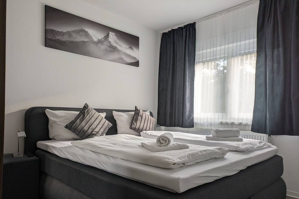 Une chambre d’hôtel moderne avec un grand lit, des rideaux foncés, un tableau représentant un paysage de montagne et une table de chevet avec une lampe.