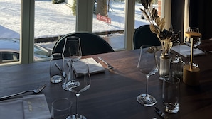 Een eettafel met wijnglazen en bestek, een vaas met gedroogde bladeren, en een besneeerd landschap zichtbaar door grote ramen.