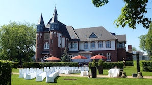 Een groot, historisch gebouw met een symmetrische gevel, meerdere torens en een verzorgd gazon met rijen witte stoelen en tafels, wat wijst op een evenement of bijeenkomst buiten.
