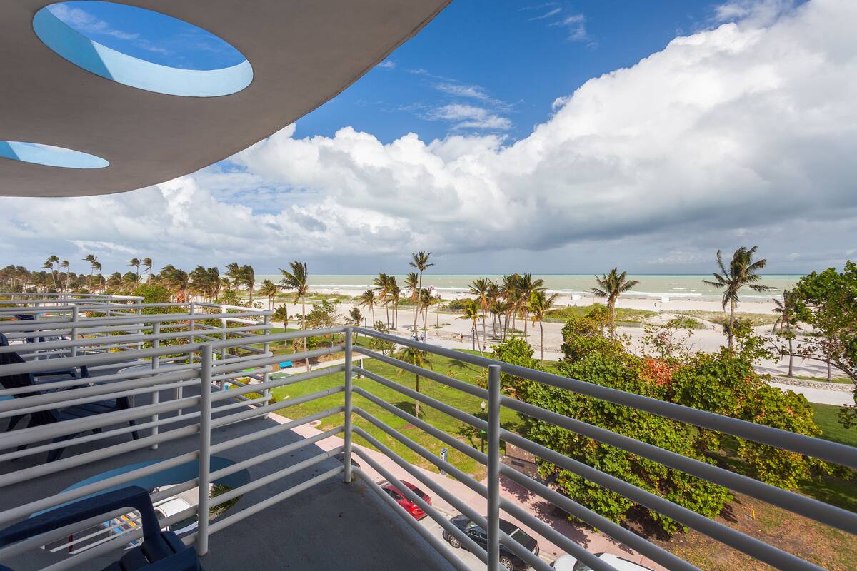 A balcony with a view of a beach, palm trees, and a cloudy sky.