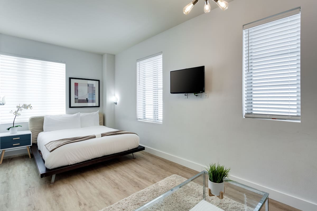 A modern bedroom with a bed, bedside tables, a TV, and large windows with blinds.