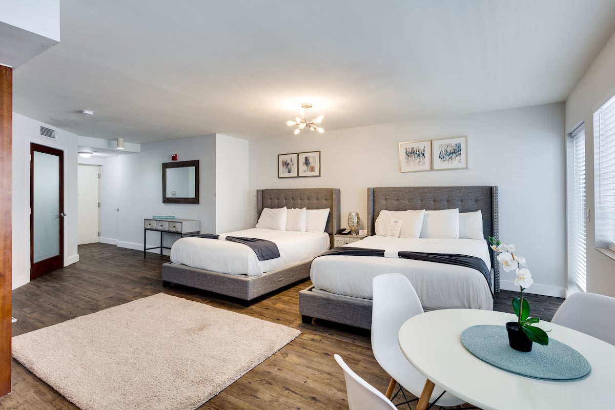 A modern hotel room with two beds, a small desk, a chair, and a dining table.