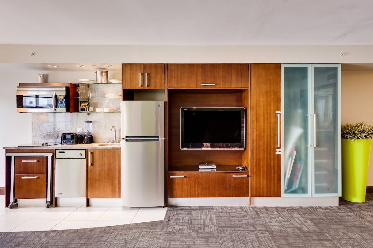 A modern kitchen with wooden cabinets, a built-in microwave, a refrigerator, and a large flat-screen TV.