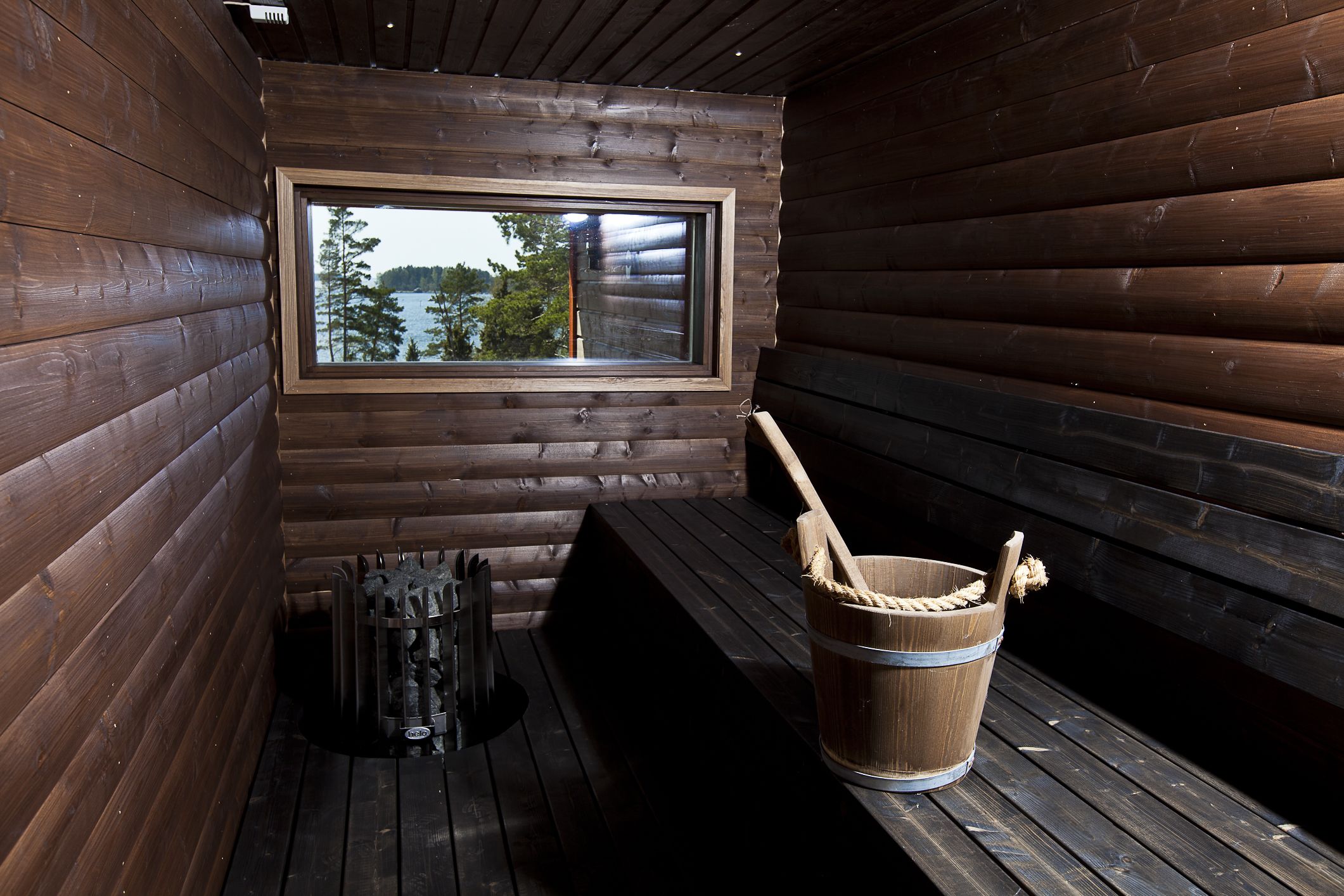 Une cabane en bois avec une fenêtre donnant sur un lac, un seau en bois et un radiateur en métal posé sur le sol.