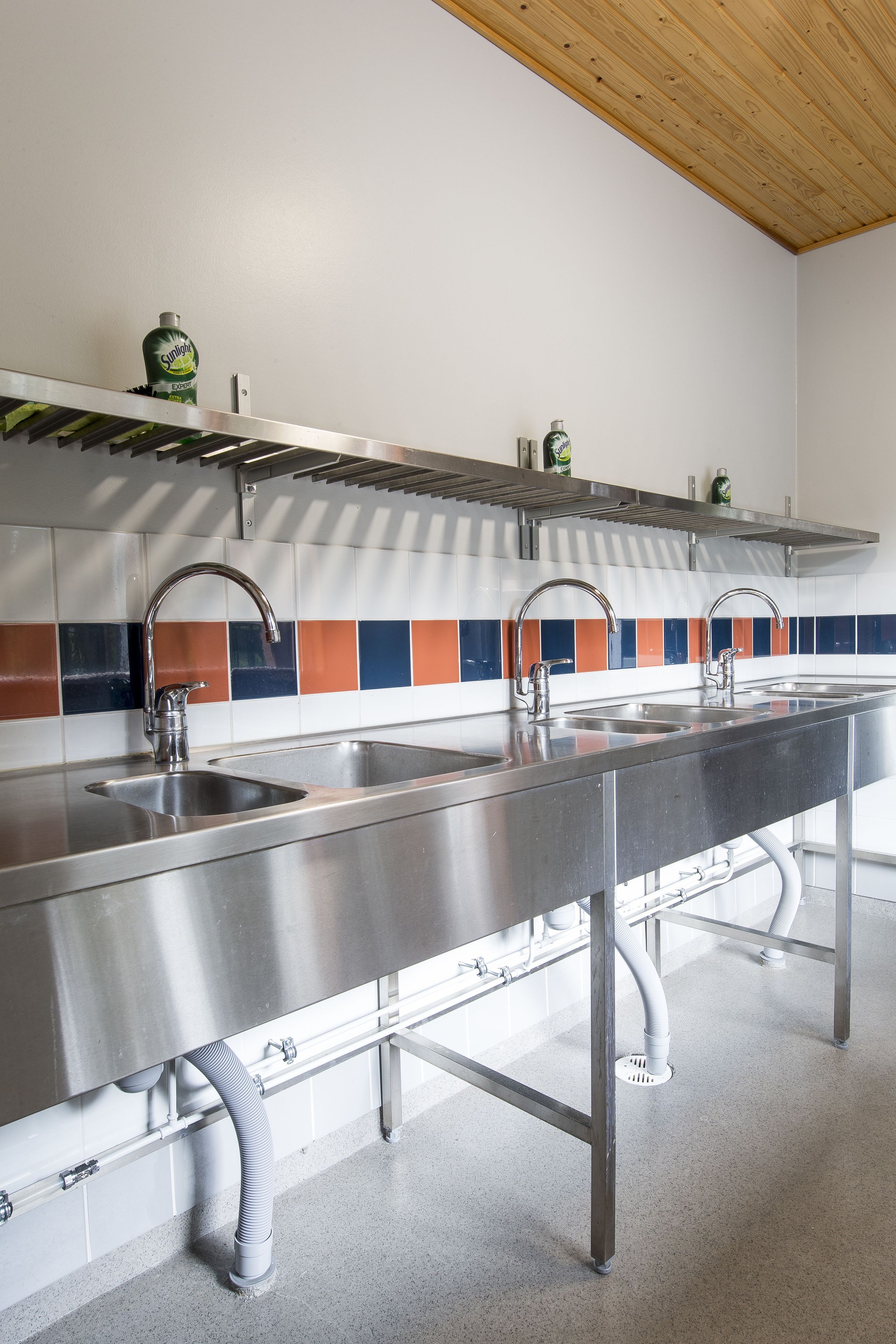 A commercial kitchen with stainless steel sinks, a tiled backsplash, and overhead ventilation.
