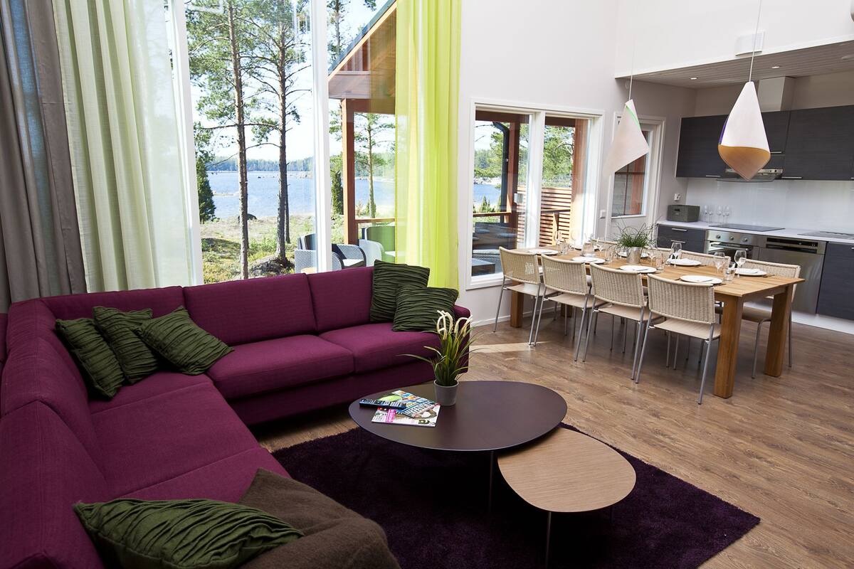 Uma sala de estar moderna com um sofá roxo, um espaço de jantar com mesa de madeira e uma cozinha com armários escuros.