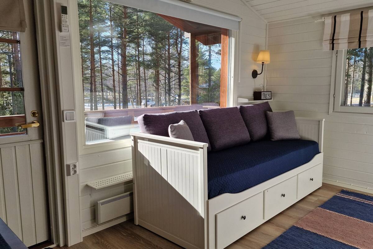 Quarto aconchegante com cama embutida, janela com vista para árvores, luminária e radiador.