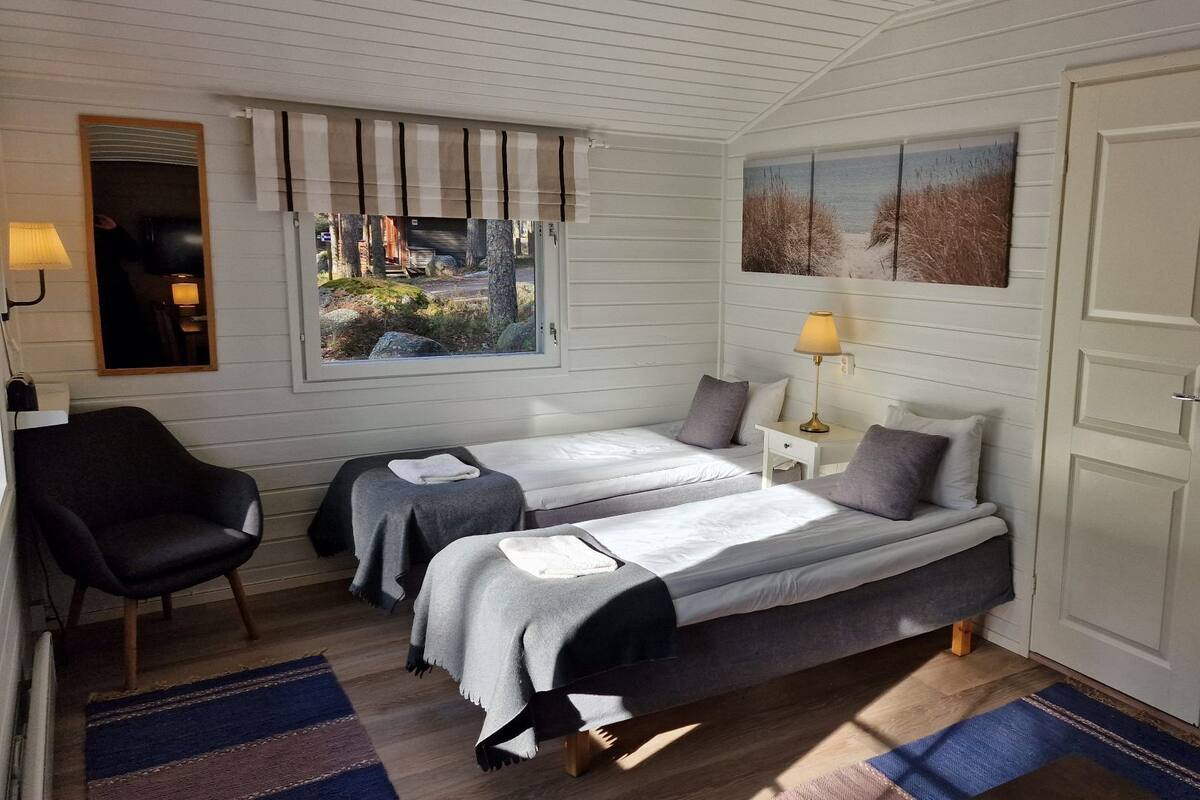 Quarto com duas camas, uma cadeira, janela com vista, porta e uma pequena mesa com abajur.