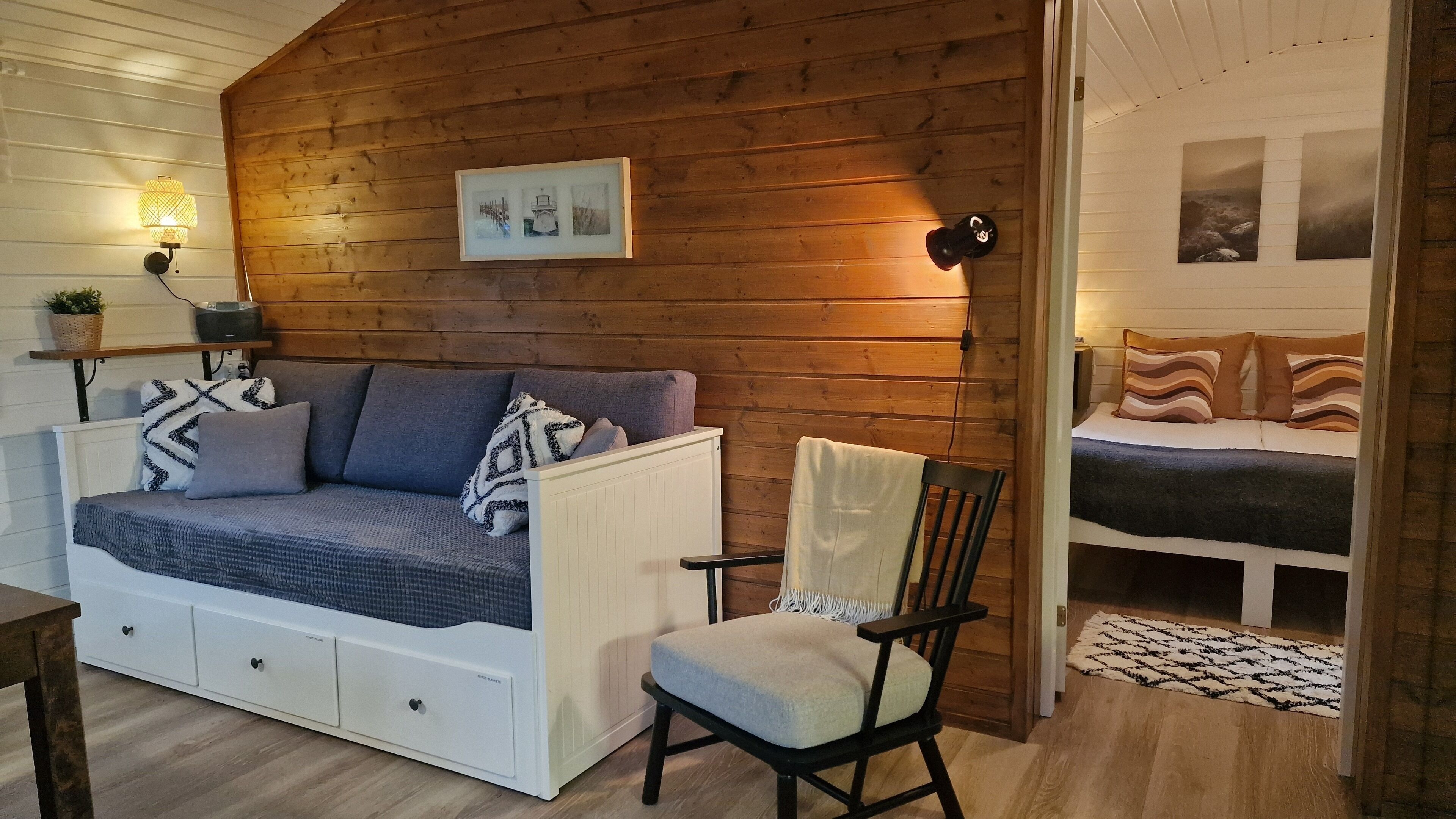 A cozy room with a wooden wall, a sofa with storage drawers, a chair, and a bed with striped pillows.