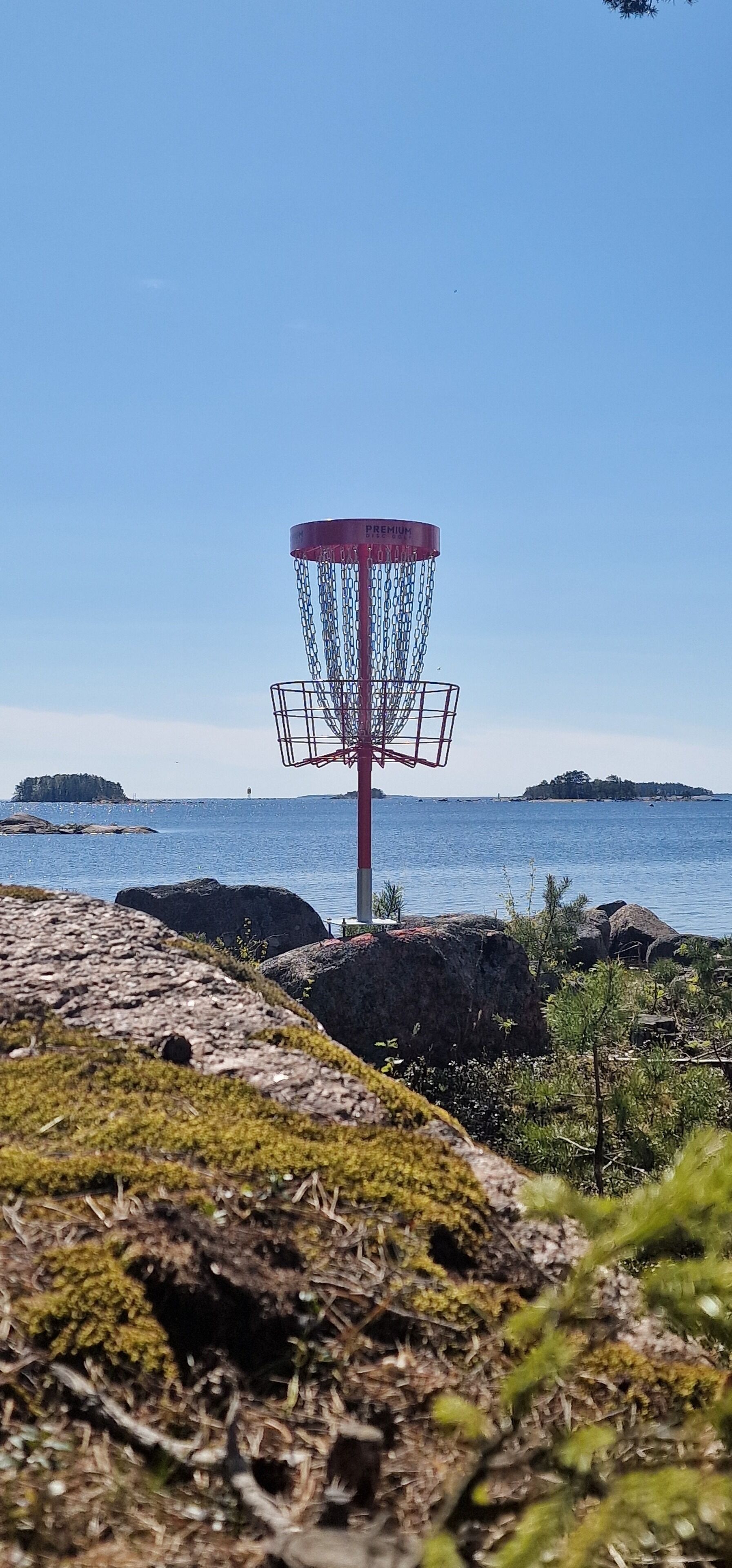 Un cesto de disc golf con una jaula de malla de cadena, sobre un fondo de cielo azul despejado y agua tranquila.