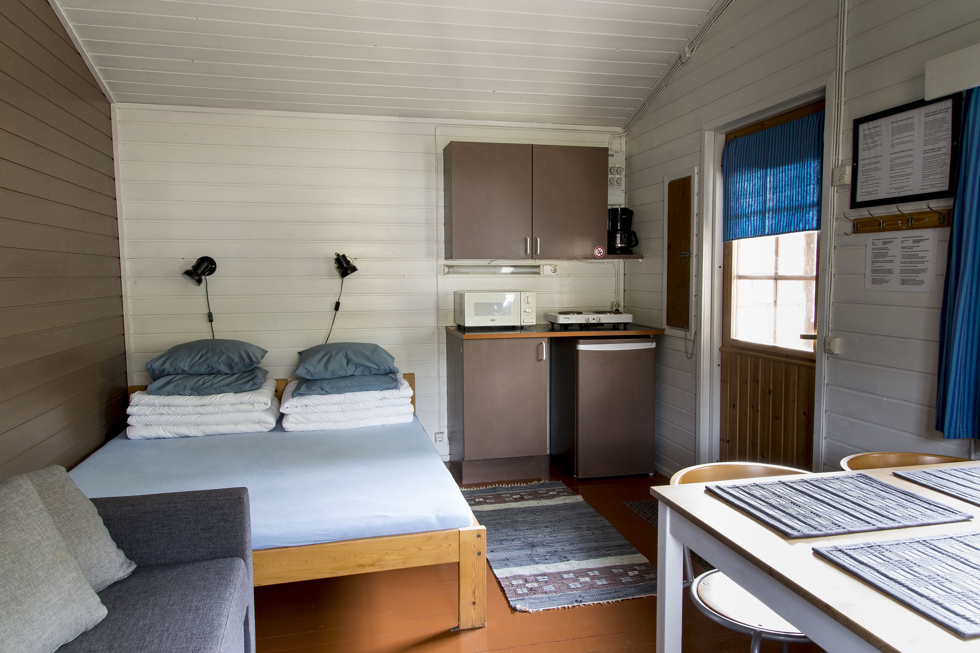 Ein kleiner, kompakter Raum mit Bett, Schreibtisch, Küchenzeile und einem mit Vorhängen versehenen Fenster.