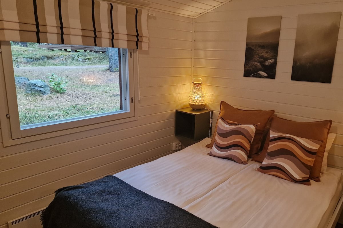 Um quarto com uma cama, duas luminárias fixadas na parede, uma janela com vista para um jardim e duas fotos emolduradas na parede.