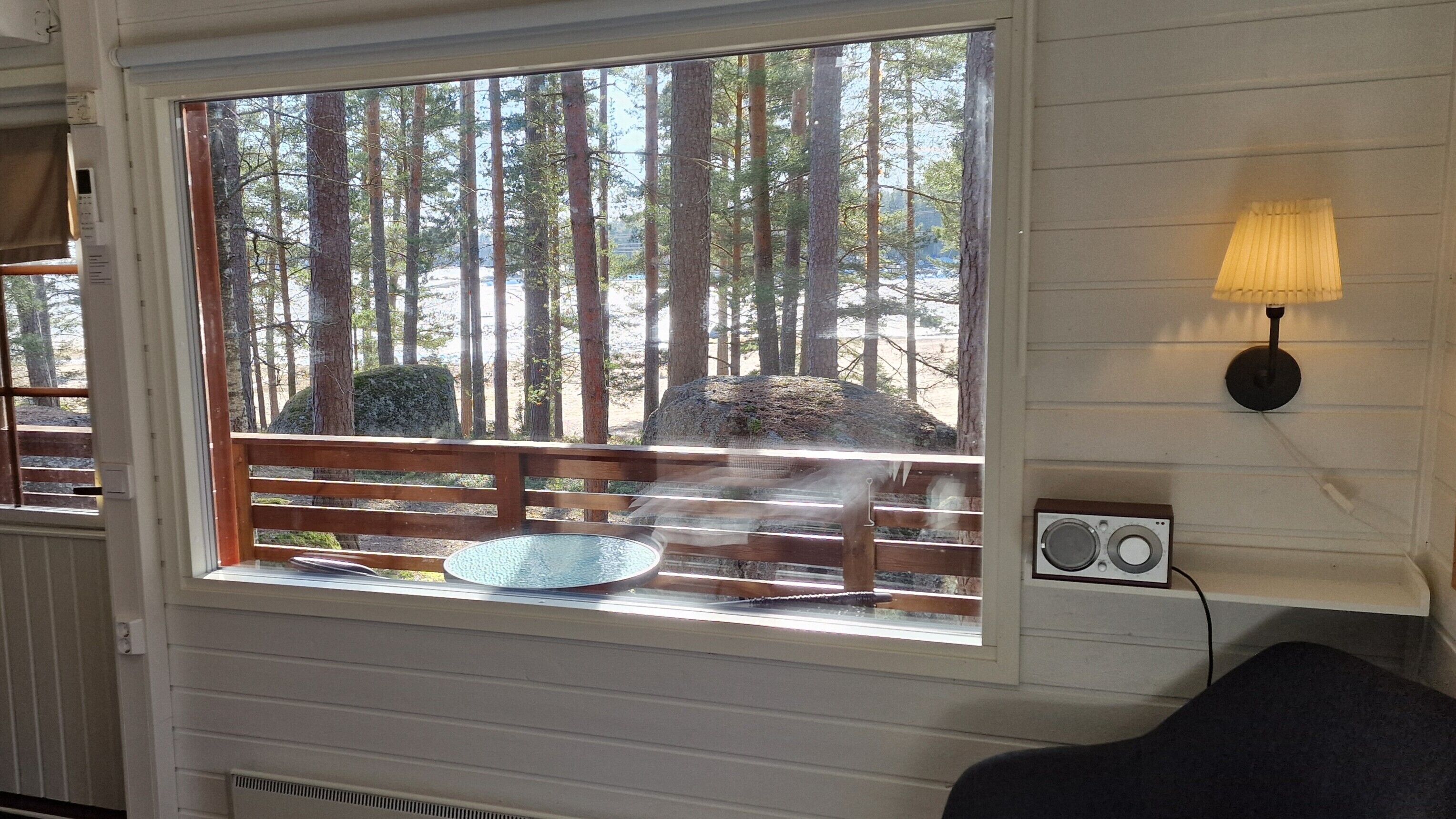 A room with a large window overlooking a forest, a wooden bench, a table with a bowl, a wall-mounted lamp, and a wall-mounted radio.