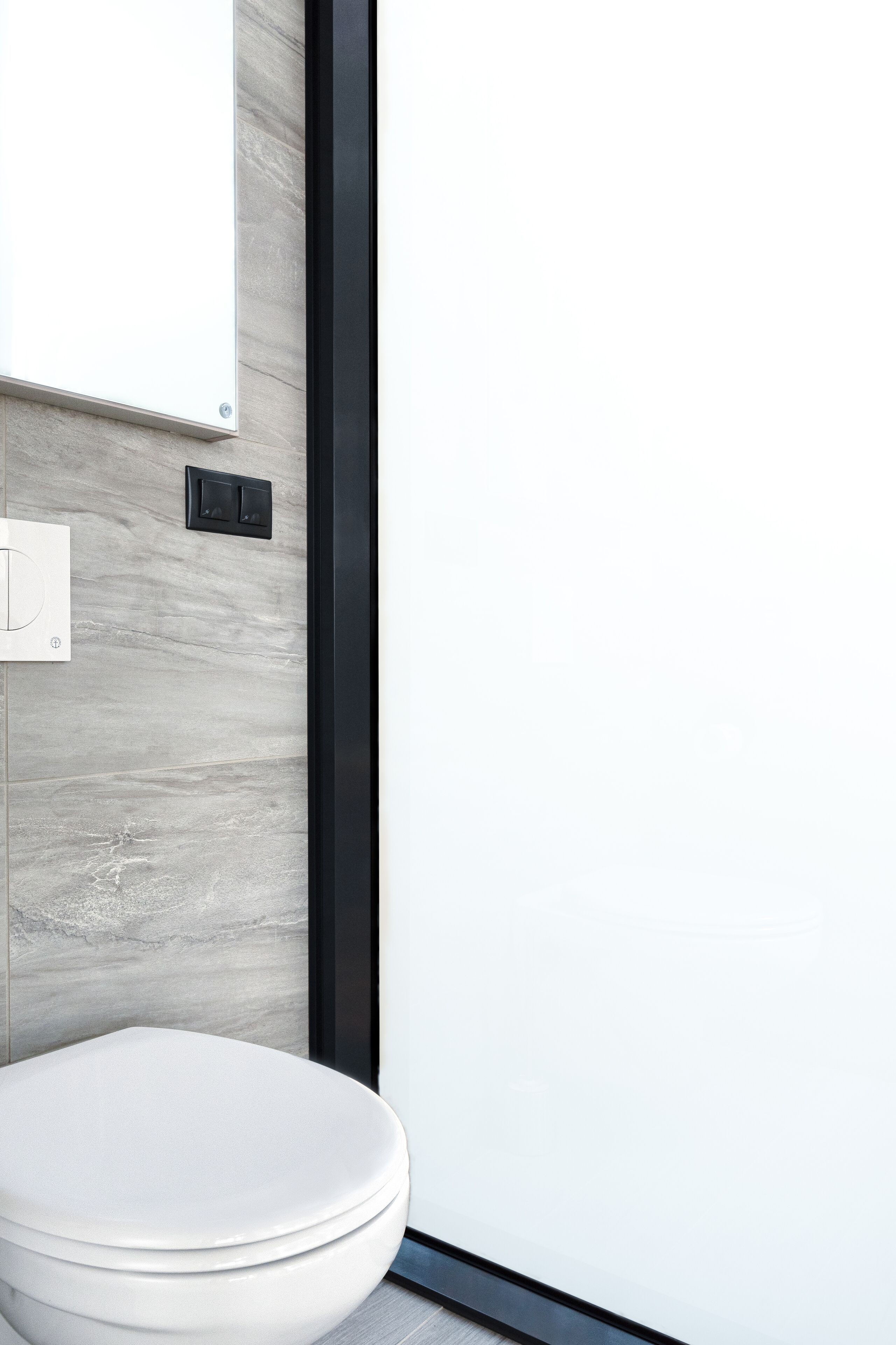 A modern bathroom with a toilet, a wall-mounted light switch, and a sleek black frame.
