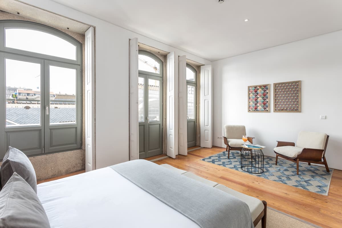 A modern bedroom with a large bed, two chairs, a small table, and a view of the city through large windows.