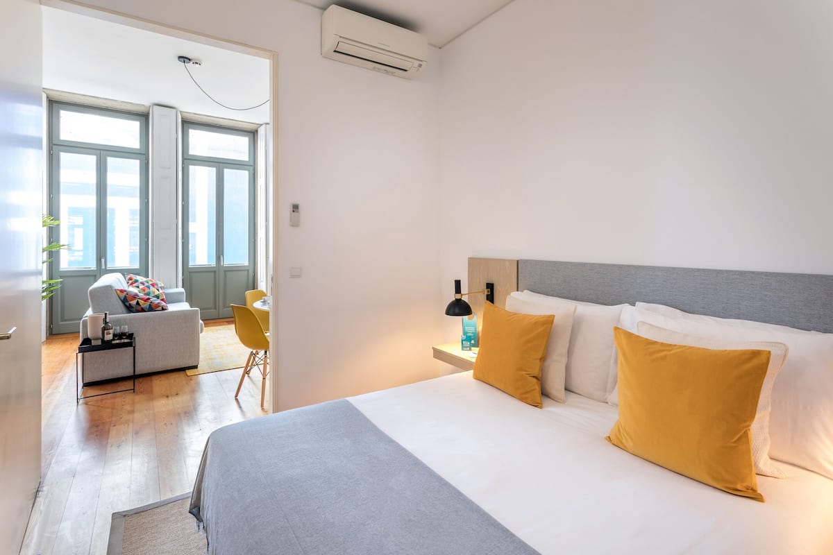 A modern hotel room with a large bed, a bedside table with a lamp, a sofa with a colorful cushion, and a view of the city through large windows.