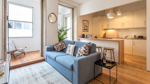 A modern living room with a blue sofa, colorful cushions, a small coffee table, and a view of a kitchen with white cabinets and pendant lights. - Villa Bolhão Apartamentos (Porto)