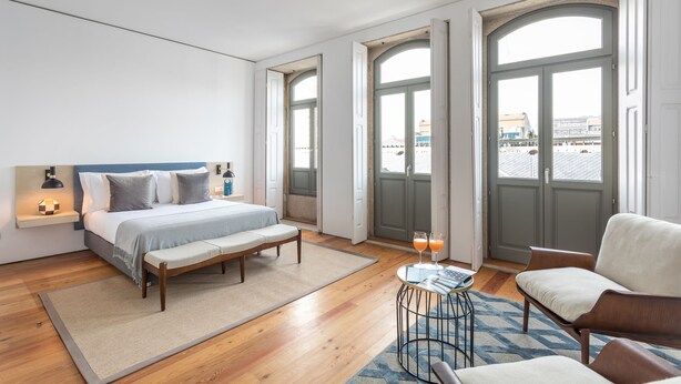 A modern hotel room with a large bed, a seating area, and a view of the city through large windows. - Villa Bolhão Apartamentos (Porto)