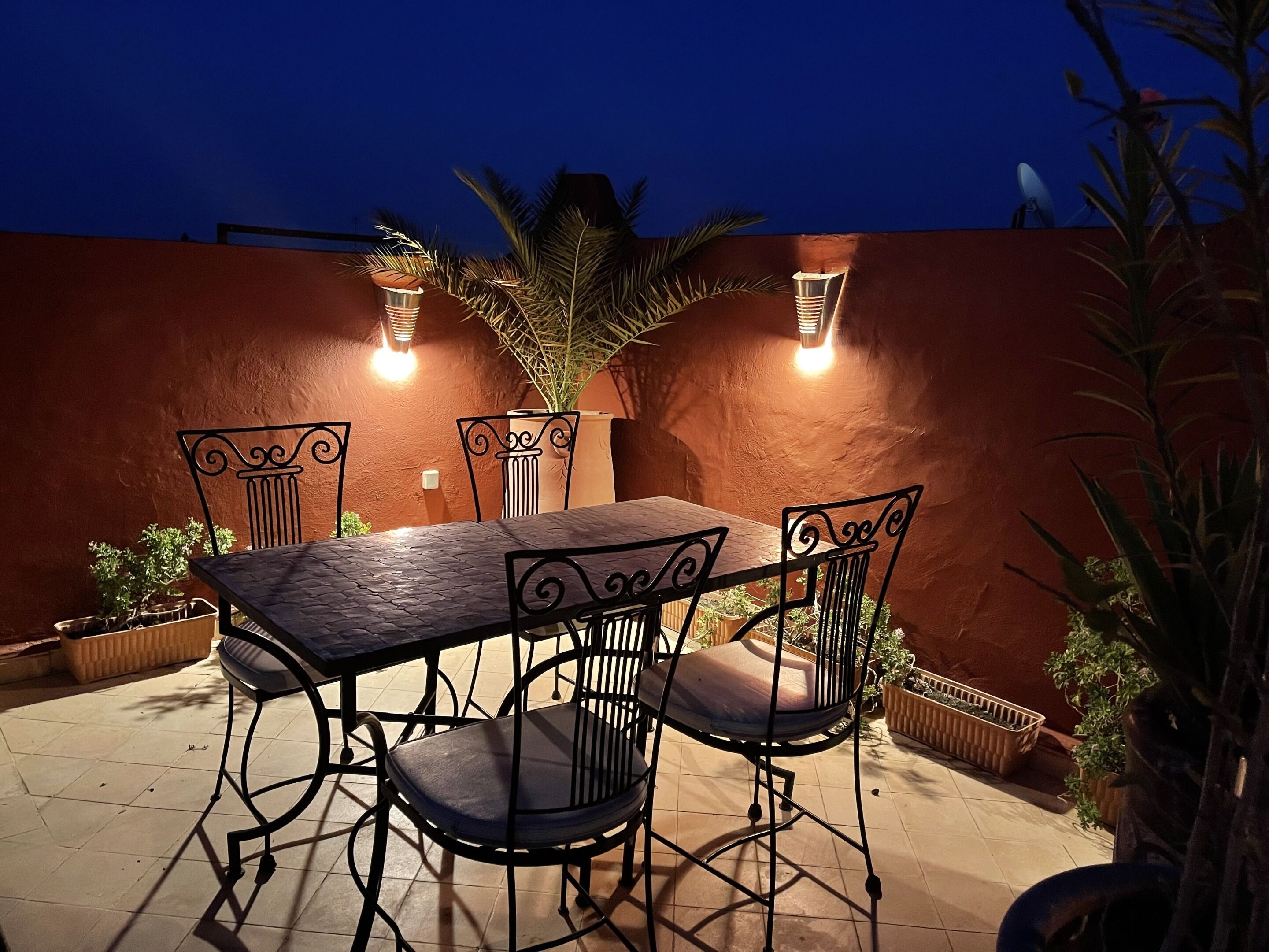 Um pátio externo com mesa e cadeiras, cercado por plantas em vasos e uma parede com textura.