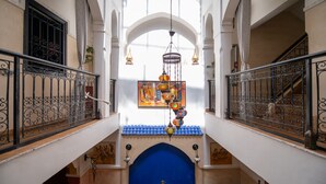 Un interior de varios niveles con barandillas metálicas ornamentales, un arco central azul y una lámpara decorativa colgante.
