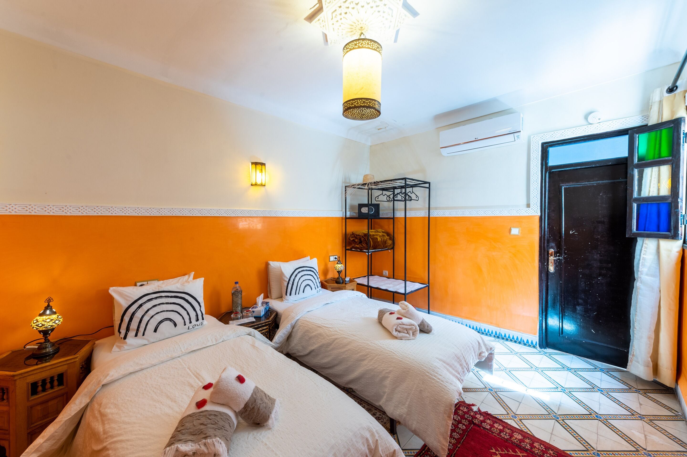 A hotel room with two beds, a small table with a lamp, a shelf with towels, and a door with a view.