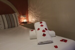 Un letto rifatto con asciugamani bianchi arrotolati e petali di rosa rossa in una camera d'albergo.
