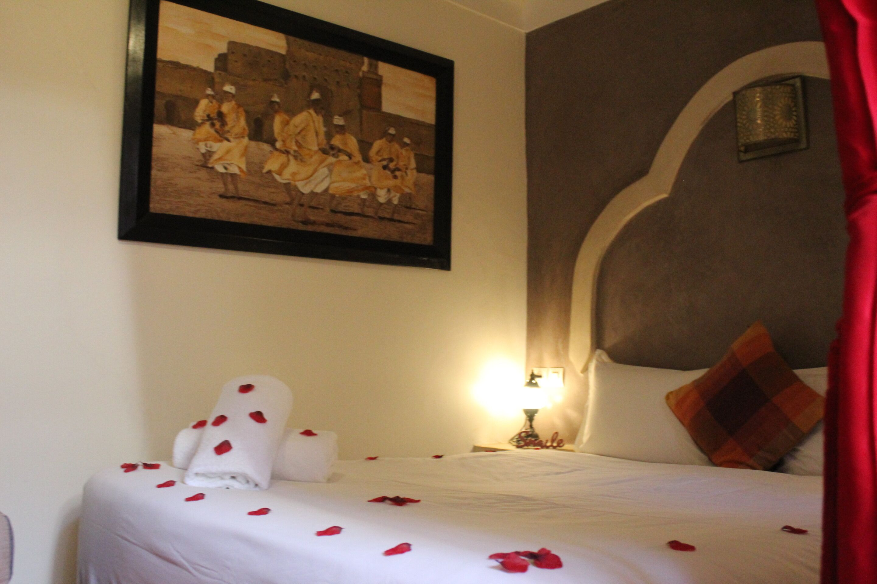 A hotel room with a bed adorned with rose petals, a framed historical painting, and a lamp on the nightstand.