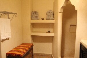 A narrow bathroom with a bench, a towel rack, and two robes hanging on a shelf.