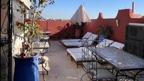 Un patio al aire libre con suelo de baldosas, mesas y sillas de metal, una planta en maceta y un cielo azul despejado.