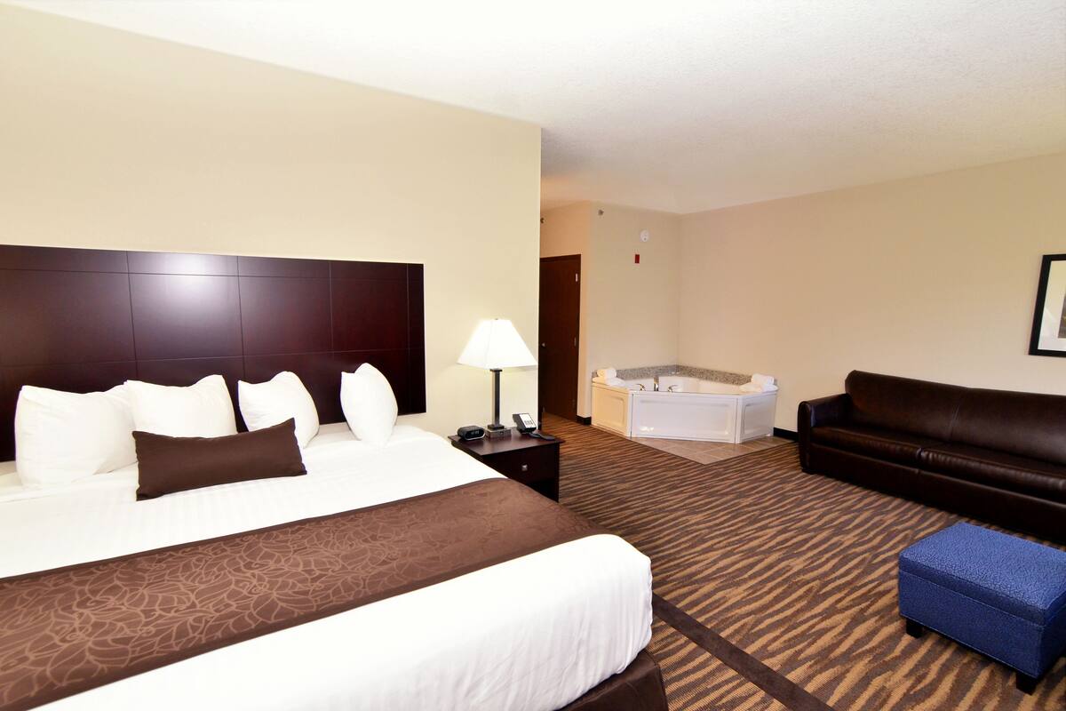 A hotel room with a large bed, a brown headboard, a white bedside table with a lamp, a blue ottoman, a brown leather sofa, and a separate bathroom with a bathtub.