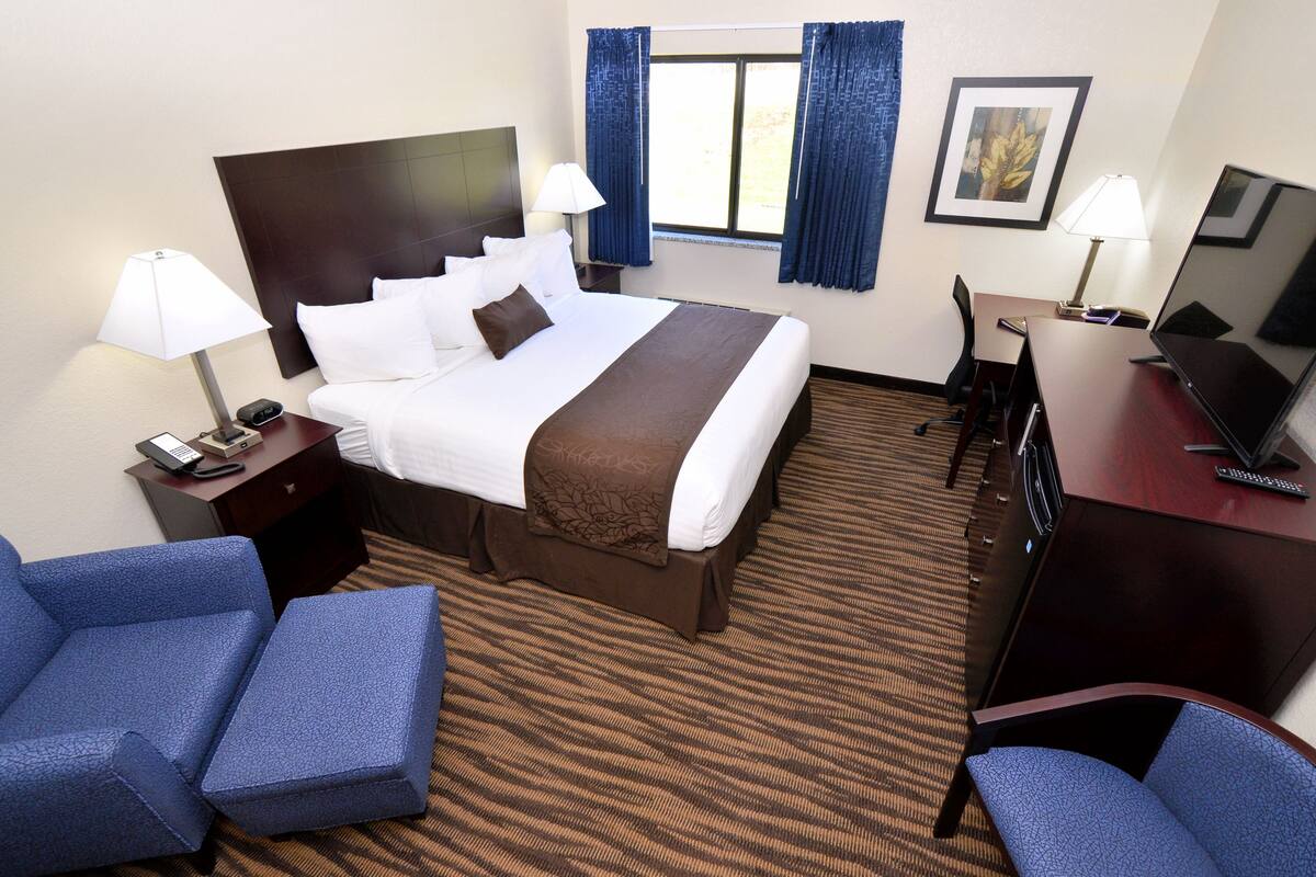 A hotel room with a large bed, two bedside tables with lamps, a desk with a computer, and two blue armchairs.