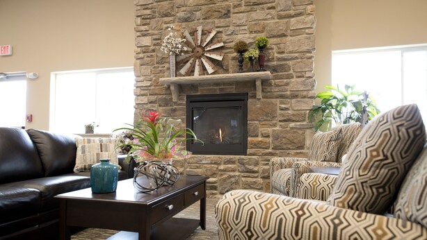 A living room with a stone fireplace, a black leather sofa, patterned armchairs, and a coffee table with decorative items. - Cornerstone Inn & Suites (Oelwein)