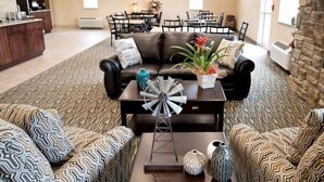 A living room with a leather sofa, patterned cushions, a coffee table with decorative items, a fireplace, and a dining area in the background. - Cornerstone Inn & Suites (Oelwein)