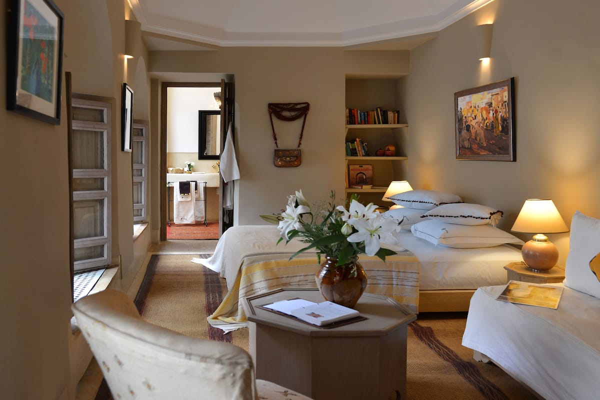 A hotel room with a bed, bedside lamps, a desk with a vase of flowers, a chair, a bookshelf, and a bathroom visible through an open door.