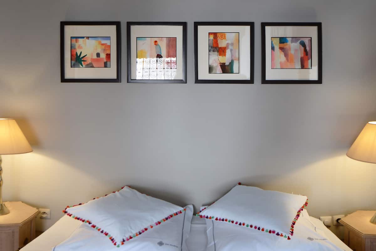 A hotel room with a bed, two bedside lamps, and four framed abstract paintings on the wall.