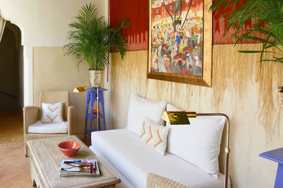 A cozy living room with a sofa, armchairs, a coffee table with magazines, a potted plant, and a vibrant painting on the wall.