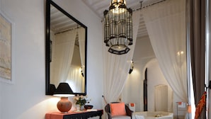 A bedroom with a bed, a nightstand, a chair, a large mirror, a chandelier, and a bathtub. - Dar Housnia (Marrakech)