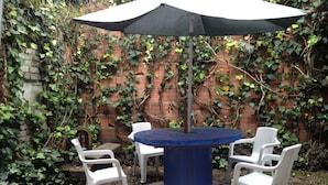 A round table with a blue top surrounded by four white plastic chairs, set against a backdrop of a brick wall partially covered with green ivy. - Karuss Hostel (Bogotá)
